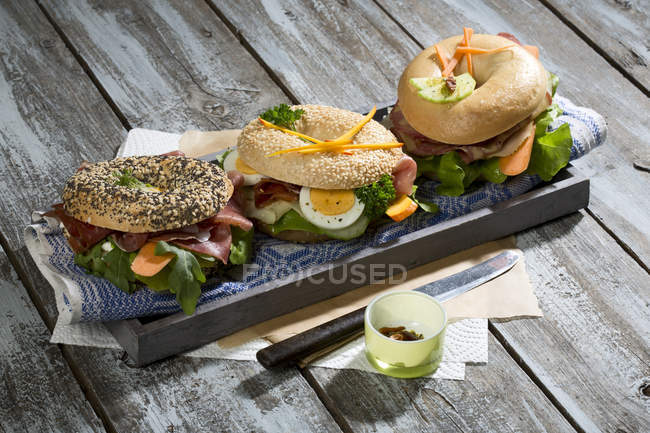 Drei verschiedene Bagels garniert mit Salami, Wurst, Speckscheiben, Rucola, Salat, Gurkenmöhre, Ei, Frischkäse und Kresse und Petersilie — Stockfoto