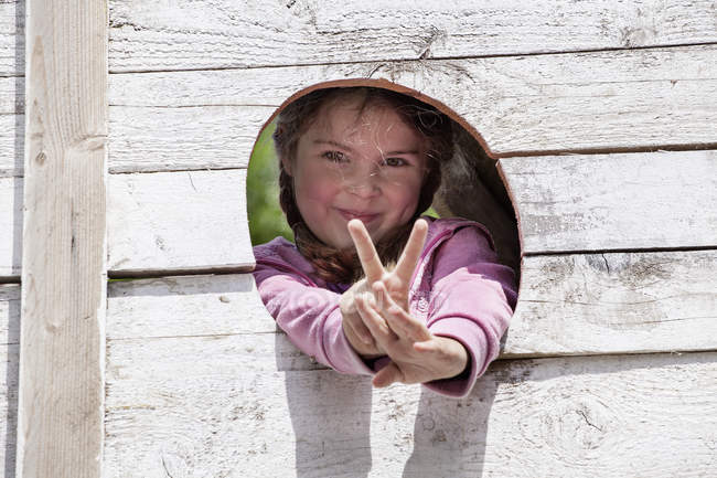 Portrait de fille montrant le signe V, souriant à la caméra dans un trou mural en bois — Photo de stock