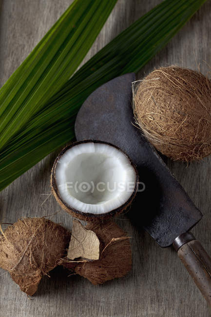 Cocos enteros y cortados a la mitad en madera oscura con cuchillo viejo y hoja - foto de stock