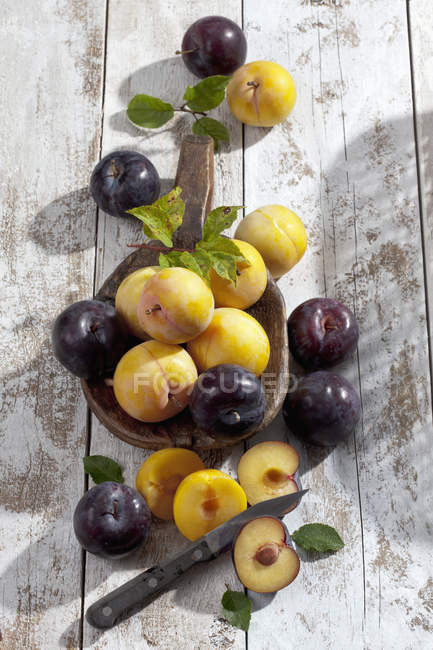 Halved and whole yellow and black plums on wooden spoon on white wooden table — Stock Photo