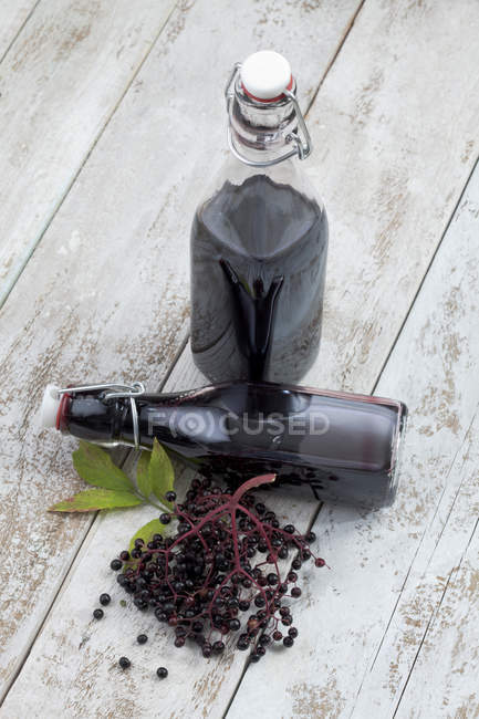 Bayas de saúco frescas y dos botellas de jugo de saúco sobre una mesa de madera blanca - foto de stock