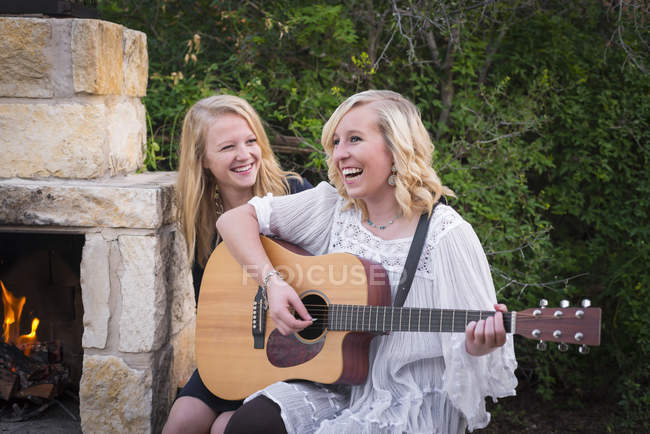 Dos niñas adolescentes felices con la guitarra sentada al lado de la chimenea - foto de stock