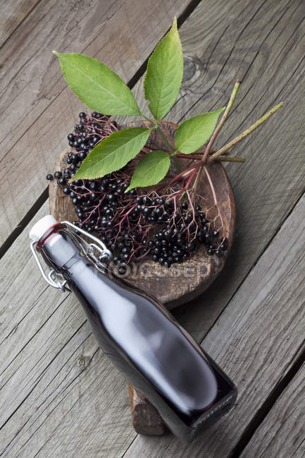 Elderberries with leaves in wooden bowl with bottle of elderberry juice on dark wood — Stock Photo