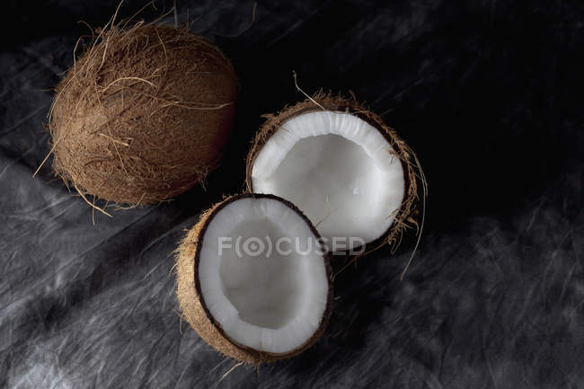 Cocos frescos enteros y cortados a la mitad sobre textil negro - foto de stock