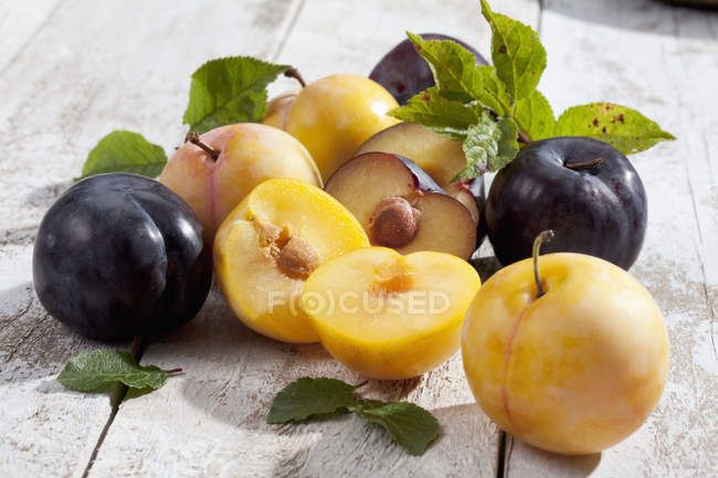 Halved and whole yellow and black plums on white wood with leaves — Stock Photo