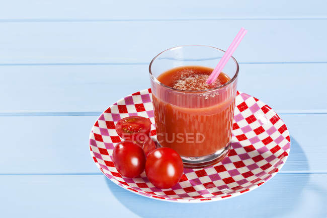 Primer plano del vaso de jugo de tomate fresco con tomates y beber paja en el plato comprobado. Fondo de mesa de madera azul - foto de stock