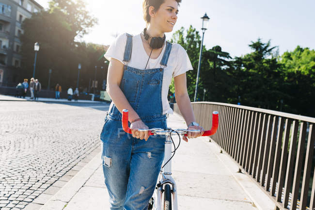 Усміхнена молода жінка штовхає велосипед на міст — стокове фото