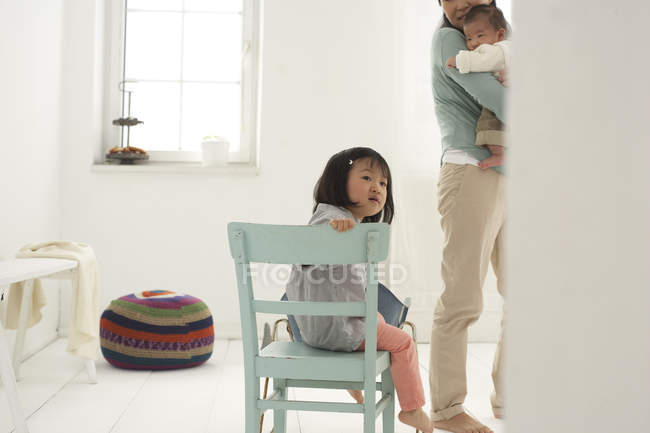 Petite fille asiatique assise sur une chaise, mère et sœur en arrière-plan — Photo de stock