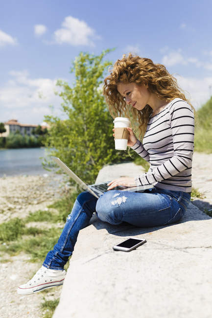 Femme avec café pour aller à l'aide d'un ordinateur portable au bord de la rivière — Photo de stock