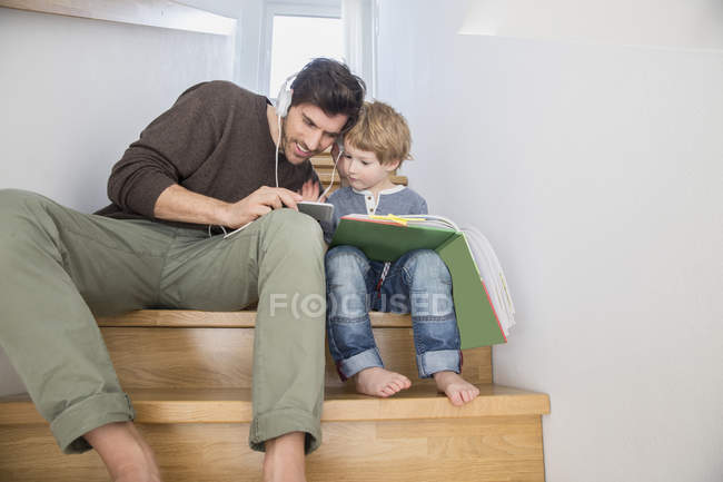 Padre e hijo sentados en las escaleras, leyendo libros y escuchando música  — Niño pequeño, escuchar música - Stock Photo | #181910106