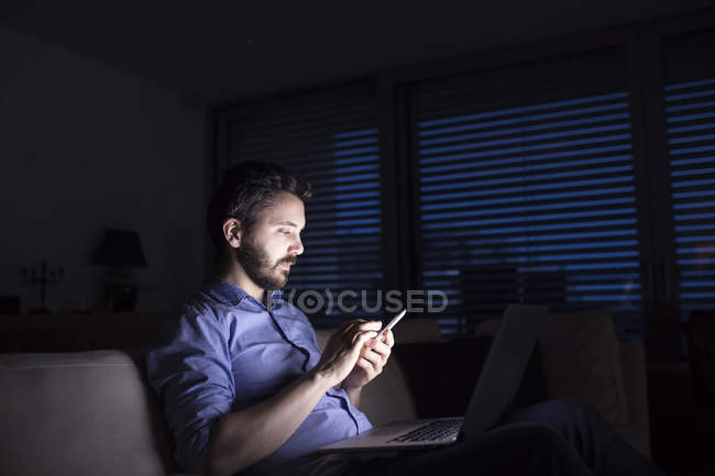 Hombre que trabaja de noche usando un teléfono inteligente y un portátil - foto de stock