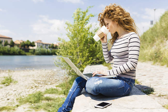 Donna che beve caffè per andare durante l'utilizzo del computer portatile a riva del fiume — Foto stock