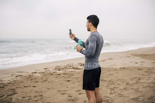 Спортивный молодой человек, пользующийся сотовым телефоном и держащий бутылку на пляже — стоковое фото