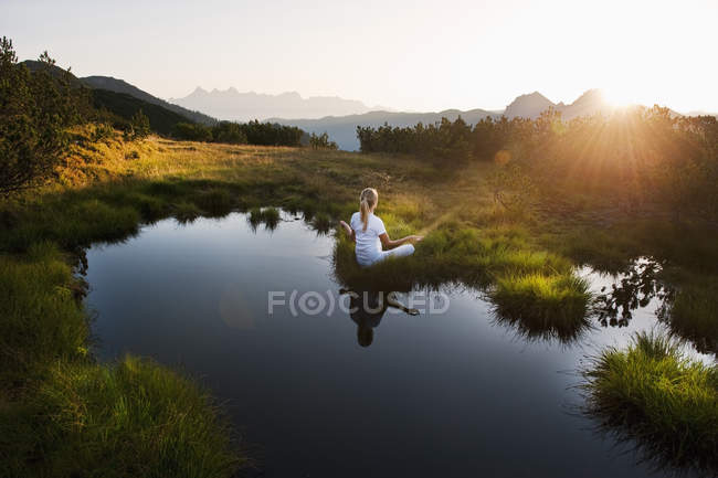 Áustria, Condado de Salzburgo, Jovem mulher sentada perto do lago de montanha e fazendo meditação — Fotografia de Stock