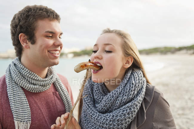 Young man feeding grilled sausage to teenage girl — Stock Photo
