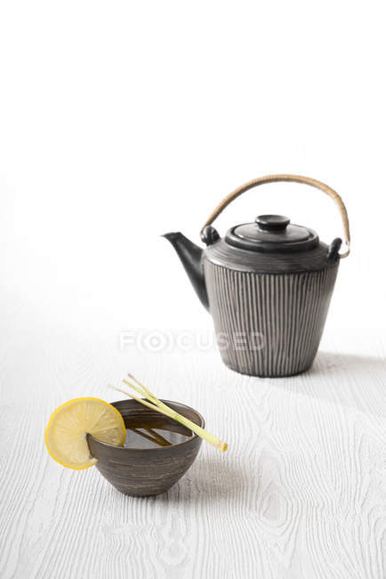 Té Sencha verde en un tazón de té con hierba de limón, una tetera sobre la superficie blanca - foto de stock