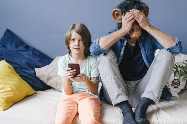 Father wearing monkey mask sitting next to son using smartphone at home — Stock Photo