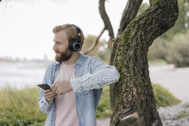 Красивий бородатий людина з смартфоном і навушниками прослуховування музики на відкритому повітрі на дереві в природі — Stock Photo