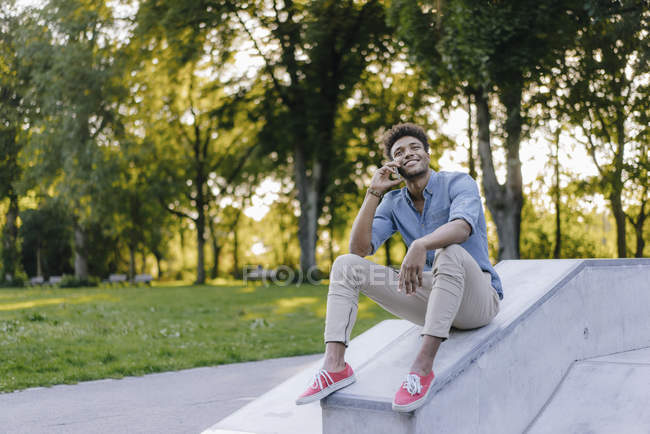 Усміхнений молодий афроамериканець чоловік розмовляє стільникового телефону, сидячи в скейтпарк — стокове фото