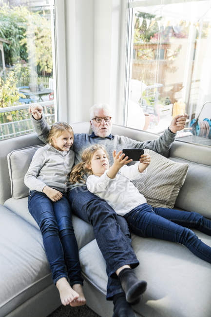 Две девочки и дедушка на диване делают селфи — стоковое фото