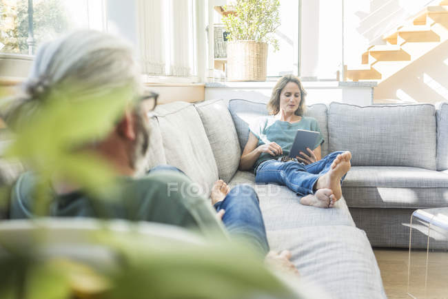 Pareja madura relajándose en el sofá en casa con la mujer sosteniendo la tableta - foto de stock