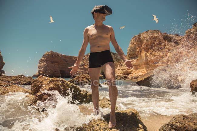 Homem maduro vestindo óculos VR entre rochas na praia — Fotografia de Stock