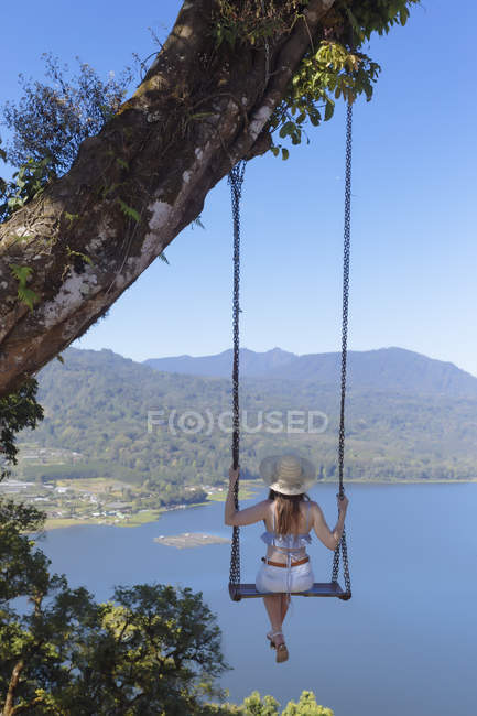 Indonesien, bali, junge Frau sitzt auf einer Schaukel unter einem Baum mit Blick auf die Küstenlandschaft — Stockfoto