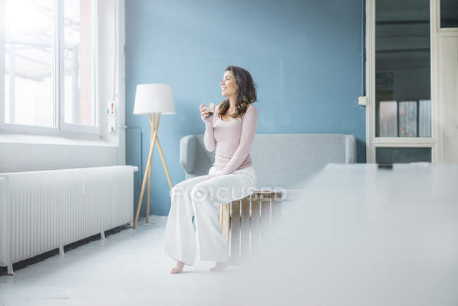 Jeune femme relaxante avec un verre de café dans le grenier et regardant par la fenêtre — Photo de stock