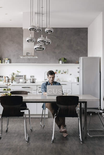 Young man sitting at table in the kitchen and working on laptop — Stock Photo