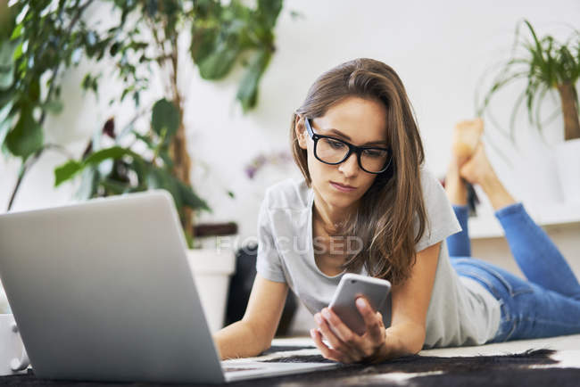 Young woman at home lying on the floor with cell phone and laptop — Stock Photo