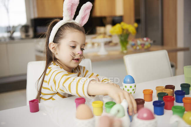 Chica con orejas de conejo sentado a la mesa y pintando huevos de Pascua - foto de stock