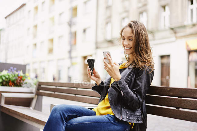 Portrait of happy woman sitting on bench with ice cream cone and looking at cell phone — Stock Photo