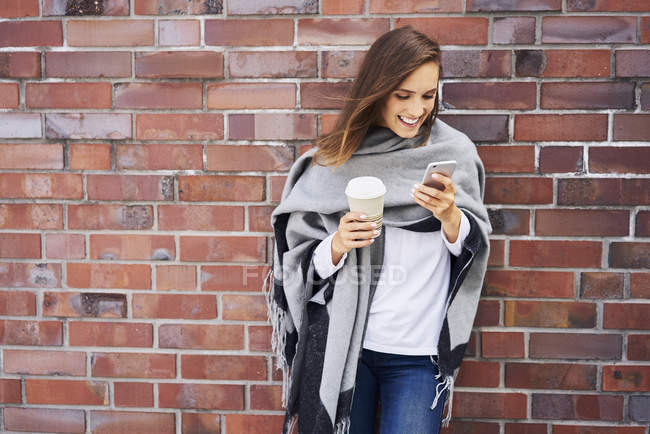 Mujer feliz con café para ir mirando el teléfono celular frente a la pared de ladrillo - foto de stock