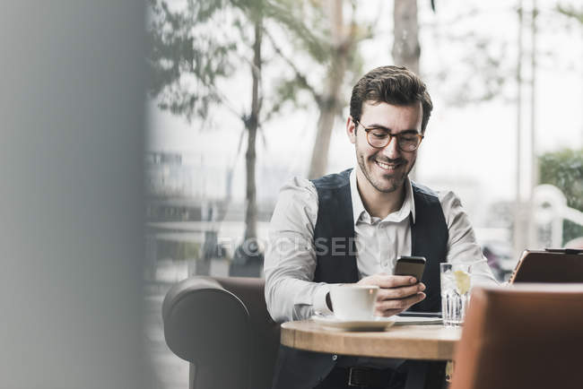 Smiling young man working in a cafe and looking at cell phone — Stock Photo