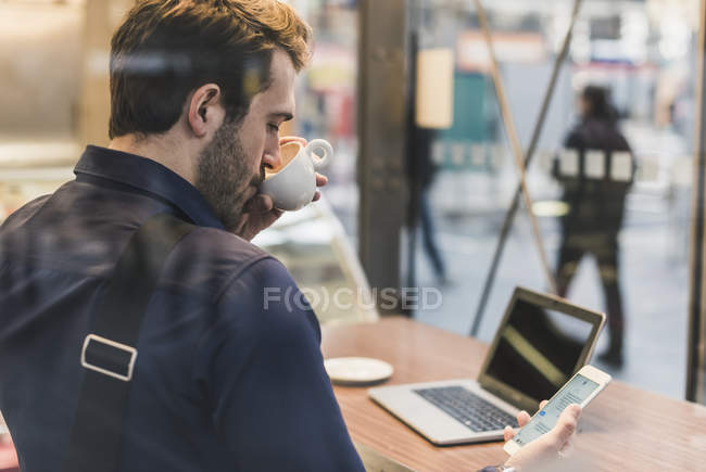 Young businessman in a cafe at train station with cell phone, drinking coffee from cup — Stock Photo