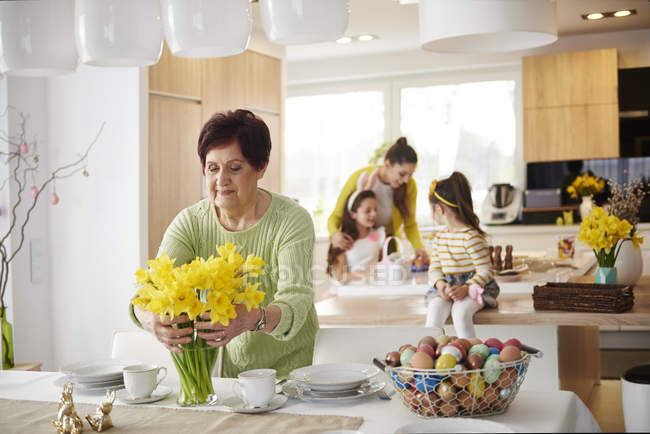 Donna anziana che organizza fiori sul tavolo da pranzo con la famiglia in background — Foto stock