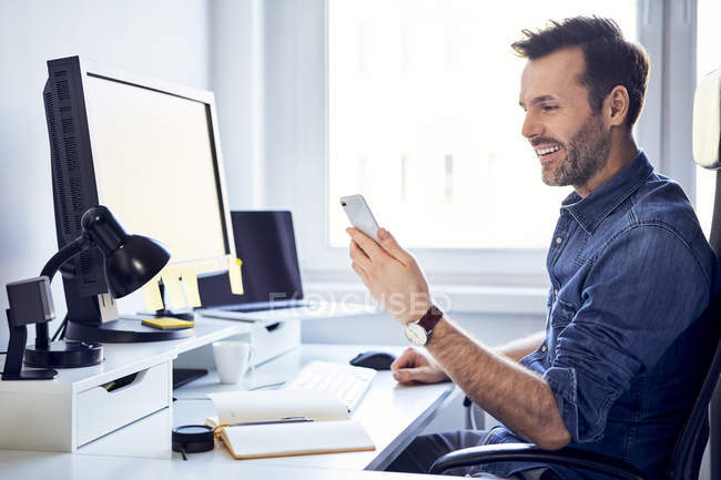Smiling man using cell phone at desk in office — Stock Photo