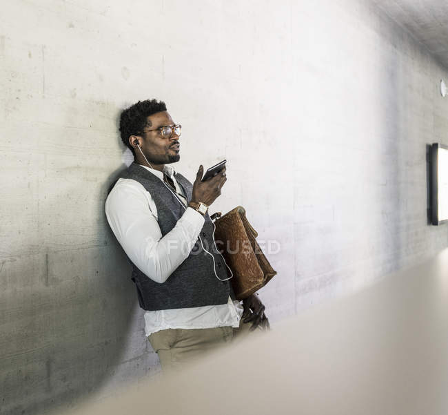 Uomo d'affari in piedi al muro di cemento con auricolari, telefono cellulare e valigetta — Foto stock