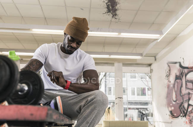 Africano americano uomo seduto su panchina in palestra e guardando su smartwatch — Foto stock