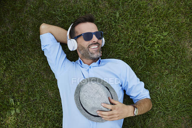 Ritratto di uomo ridente che si rilassa sul prato ascoltando musica con cuffie e cappello — Foto stock
