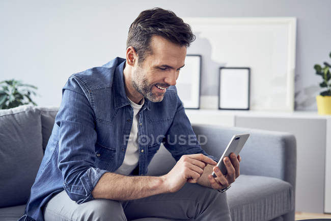 Улыбающийся мужчина сидит на диване и пользуется мобильным телефоном — стоковое фото