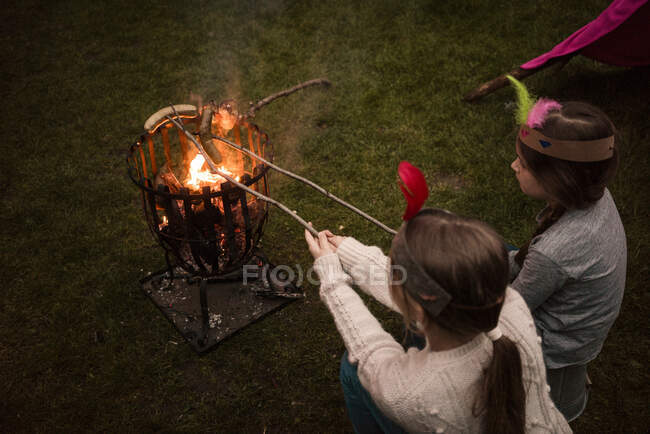 Zwei Mädchen mit Federschmuck grillen Würstchen am Lagerfeuer — Stockfoto