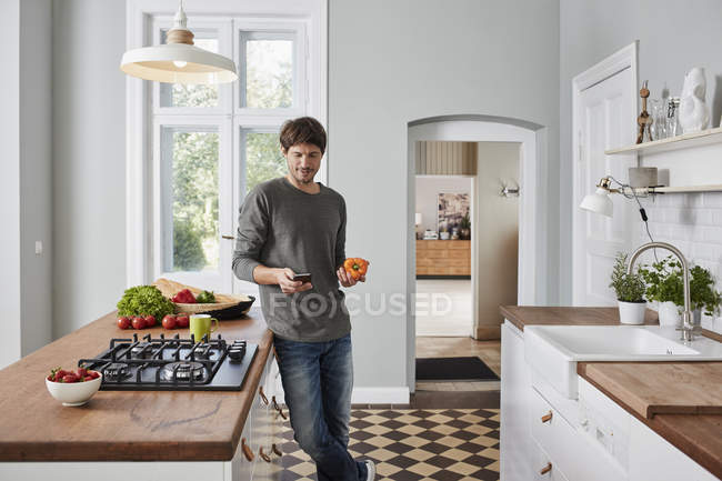 Man using smartphone and holding bell pepper in kitchen — Stock Photo