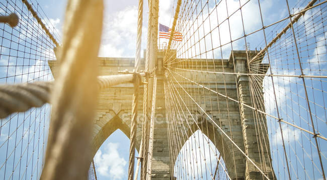 Estados Unidos, Nueva York, Brooklyn, Primer plano de los cables y arcos metálicos del puente de Brooklyn con bandera americana en la parte superior - foto de stock