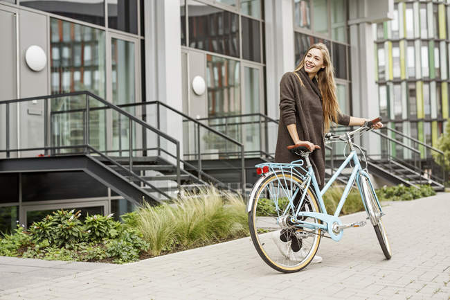 Усміхнена жінка з велосипедом стоїть перед будівлею — стокове фото