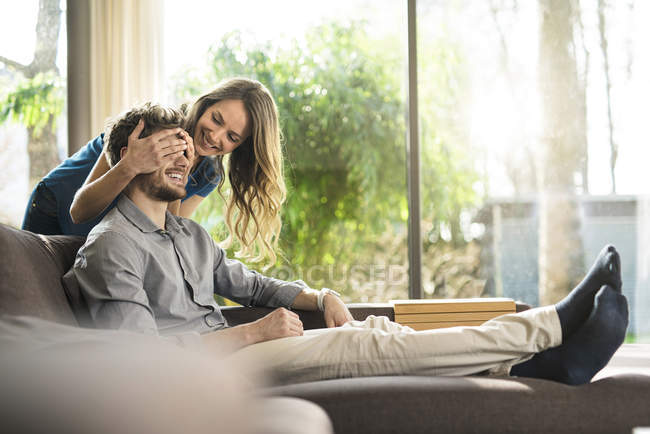 Donna sorridente che copre gli occhi del suo ragazzo sul divano di casa — Foto stock