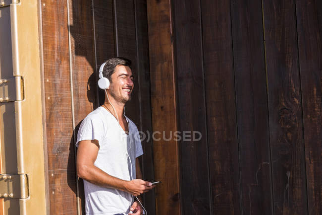 Happy young man with cell phone and headphones leaning against wooden wall — Stock Photo