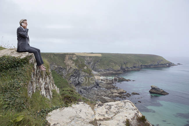 Reino Unido, Cornwall, Gwithian, hombre de negocios sentado en la costa y hablando por teléfono celular - foto de stock