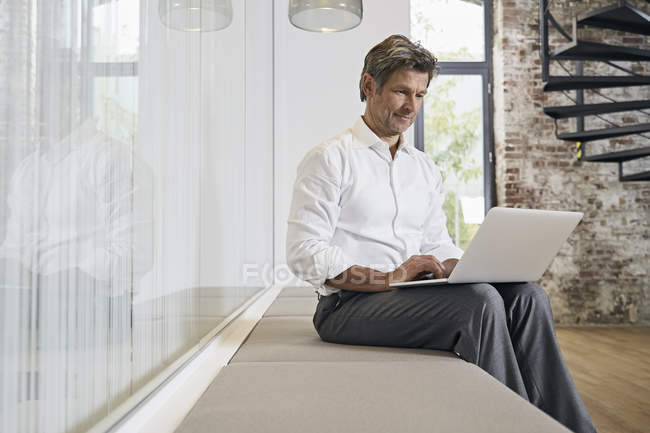 Empresário sentado no banco no escritório moderno e usando laptop — Fotografia de Stock