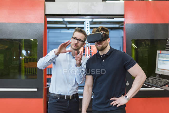 Man explaining machine to colleague wearing VR glasses in factory — Stock Photo